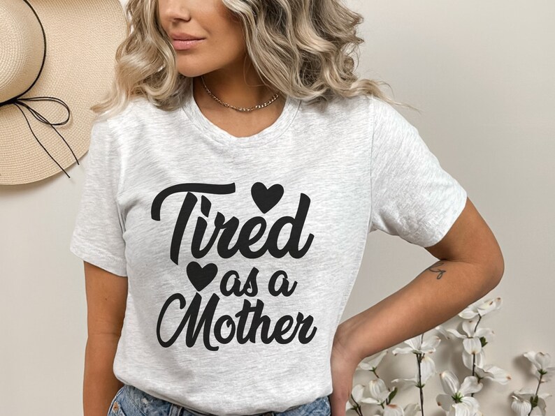 Op de afbeelding: Een grijs T-shirt met de tekst "Tired as a Mother" in zwarte cursieve letters met drie zwarte harten.
