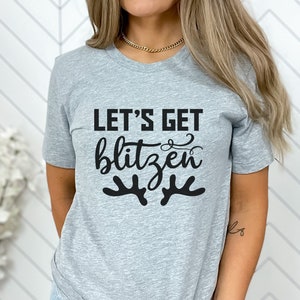 Peut inclure: Un t-shirt gris avec le texte "Let's Get Blitzen" en noir et un graphique de bois de renne stylisé.
