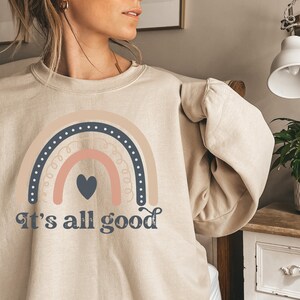 Può includere: Una felpa beige con un design a arcobaleno e il testo "It's all good" in un font retrò. L'arcobaleno è composto da strisce rosa, arancioni e blu con un cuore nero al centro.