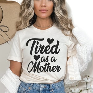 Op de afbeelding: Een wit T-shirt met de tekst "Tired as a Mother" in zwarte letters met drie zwarte hartjes. Het model draagt het shirt met een witte cardigan en een blauwe jeans.