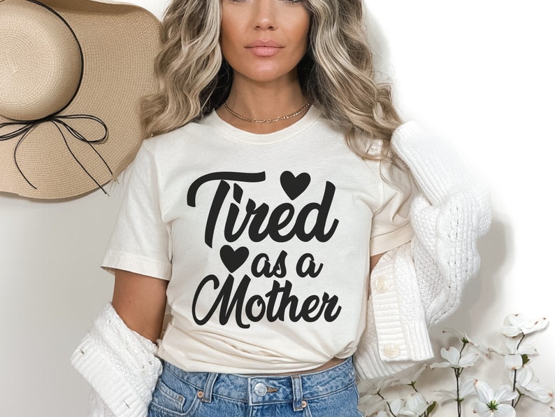 Op de afbeelding: Een wit T-shirt met de tekst "Tired as a Mother" in zwarte letters met drie zwarte hartjes. Het model draagt het shirt met een witte cardigan en een blauwe jeans.