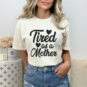 Op de afbeelding: Een wit T-shirt met de tekst "Tired as a Mother" in zwarte cursieve letters. Er zijn drie kleine zwarte hartjes boven en onder de tekst.