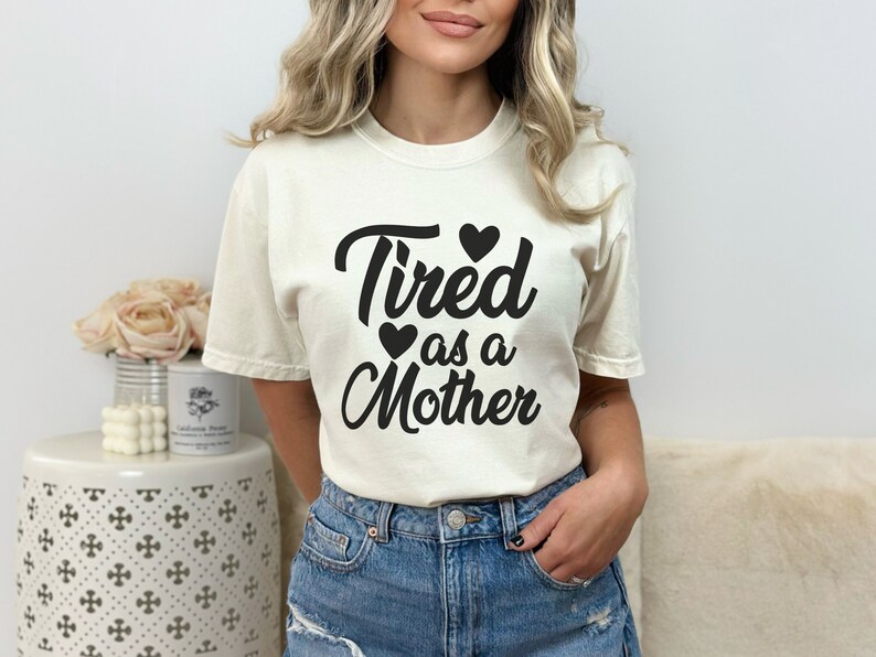 Op de afbeelding: Een wit T-shirt met de tekst "Tired as a Mother" in zwarte cursieve letters. Er zijn drie kleine zwarte hartjes boven en onder de tekst.