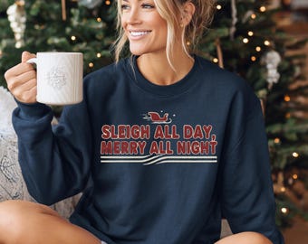 Könnte beinhalten: Marineblaues Sweatshirt mit dem Text "SLEIGH ALL DAY, MERRY ALL NIGHT" in Rot und Weiß. Das Sweatshirt zeigt eine Grafik eines Schlittens. Eine weiße Tasse wird in der Hand gehalten. Der Hintergrund umfasst einen Weihnachtsbaum.
