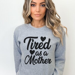 Op de afbeelding: Grijs sweatshirt met de tekst "Tired as a Mother" in zwarte cursieve letters met drie zwarte harten.