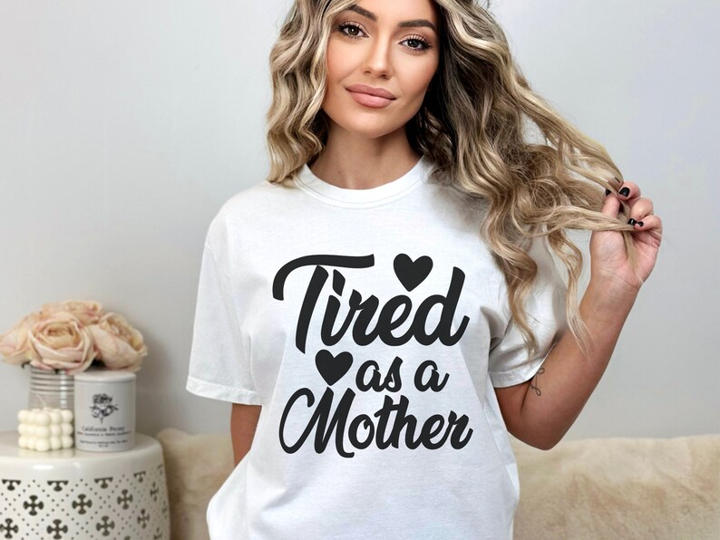 Op de afbeelding: Wit T-shirt met zwarte tekst die "Tired as a Mother" zegt met drie zwarte harten.