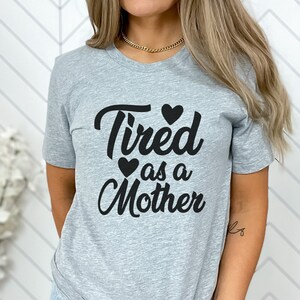 Op de afbeelding: Een grijs T-shirt met de tekst "Tired as a Mother" in zwart met twee kleine zwarte hartjes boven en onder de tekst.