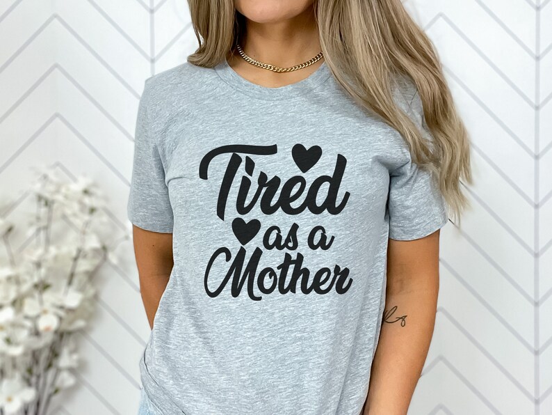 Op de afbeelding: Een grijs T-shirt met de tekst "Tired as a Mother" in zwart met twee kleine zwarte hartjes boven en onder de tekst.