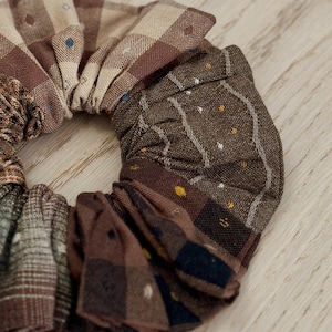 May include: A hair scrunchie constructed from multiple fabric patches in various shades of brown, tan, and dark green. The fabric features diverse patterns including plaid, stripes, and small embroidered dots in yellow, blue, and white.