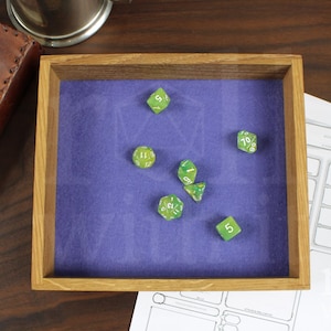 Könnte beinhalten: Würfeltablett aus Holz mit lila Filzinnenraum, das mehrere grüne Würfel enthält. Ein ledergebundenes Notizbuch und ein Bleistift mit Charakterbögen sind ebenfalls sichtbar, was auf ein Tabletop-Gaming-Thema hindeutet.