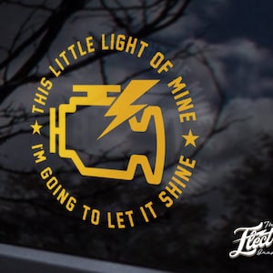 Könnte beinhalten: Ein gelber runder Aufkleber mit dem Text "This little light of mine, I'm going to let it shine" und einem stilisierten Bild einer Auto-Kontrollleuchte. Der Aufkleber befindet sich auf einer schwarzen Autoscheibe.