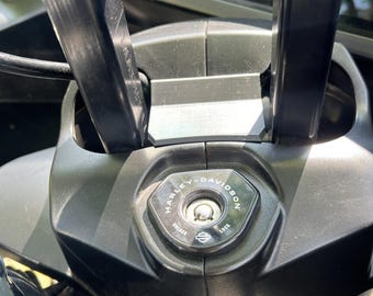 Puede incluir: Primer plano del interruptor de encendido de una motocicleta Harley-Davidson. El interruptor es negro con el logotipo de Harley-Davidson y las palabras "UNLOCK" y "LOCK". El encendido está rodeado de manillares negros y un salpicadero negro.
