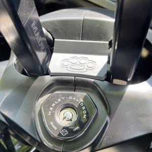 May include: Close-up of a black motorcycle ignition switch with the Harley-Davidson logo. The ignition is surrounded by black metal components, including a bracket with a brass knuckle design and a bar that says "MADE IN USA".