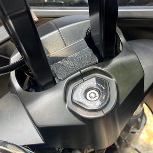 May include: Close-up of a motorcycle's handlebar area, featuring a Harley-Davidson ignition switch. The black components include handlebars, a console with a spiderweb design, and the ignition switch with the Harley-Davidson logo.