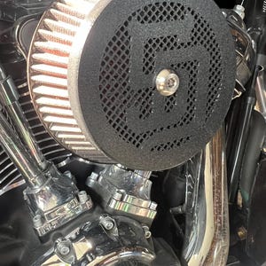 May include: Close-up of a motorcycle engine with a black air filter cover featuring a geometric design. The engine components are chrome and reflect light, with the Harley-Davidson logo visible.