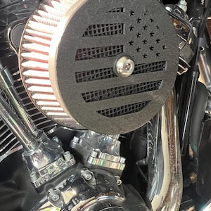 May include: A motorcycle engine featuring a custom air filter with an American flag design. The filter is black, and the engine components are chrome. The image highlights the contrast between the black filter and the shiny metal parts, showcasing a custom modification.