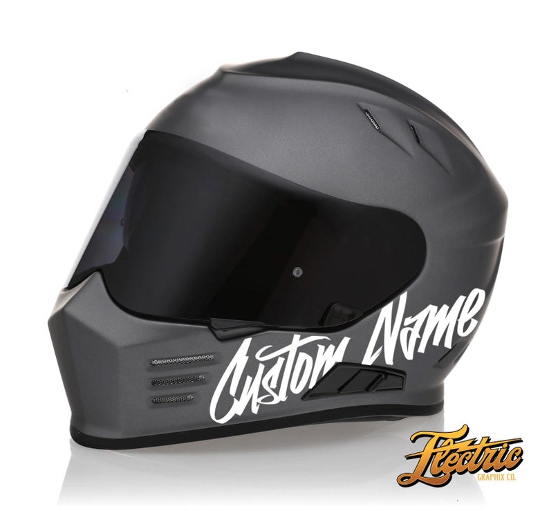 May include: A gray motorcycle helmet with a white custom name decal. The decal is in a cursive font and says "Custom Name".