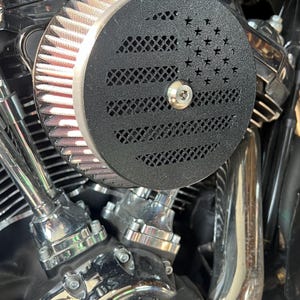 May include: Close-up of a motorcycle air filter with a black cover featuring a star and stripe design. The filter is round with a mesh-like pattern and a silver bolt. The motorcycle engine components are visible.