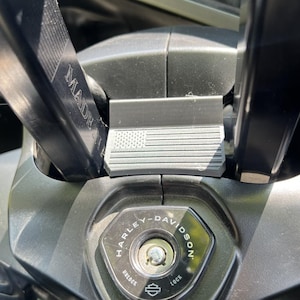 Puede incluir: Primer plano del área de encendido de una motocicleta. La ranura de la llave está rodeada por un panel negro con el logotipo de Harley-Davidson. Se ve una pequeña pieza de metal con un diseño de bandera estadounidense.