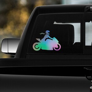 May include: A colorful iridescent decal of a person riding a motorcycle. The decal is on the back window of a black pickup truck.