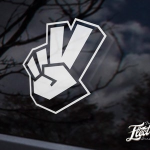 Puede incluir: Pegatina blanca con el signo de la paz con un diseño gráfico audaz. La pegatina está sobre un fondo negro.