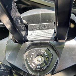 May include: Close-up of a motorcycle's ignition area. The ignition switch is surrounded by a black panel with the Harley-Davidson logo. Above the ignition is a metal plate with the text "MERICA!" and an American flag design. The handlebar has "MADE IN USA" text.