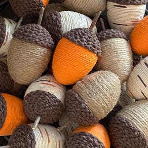 May include: A collection of handmade acorn ornaments. The acorn caps are dark brown, and the bodies are wrapped in twine or orange yarn. Some acorns have small twig stems. The acorns are a fall-themed decoration.