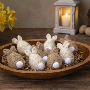 May include: A wooden bowl holds several handmade Easter bunny decorations. The bunnies are made of twine and have white pom-pom tails. The bowl sits on a wooden surface, with a vase of yellow flowers in the background.