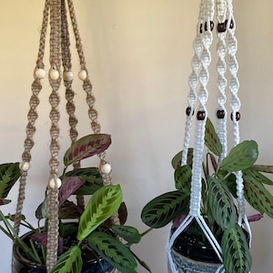 May include: Two macrame plant hangers with wooden beads. One is made of natural jute rope and the other is white cotton rope. Both are holding potted plants.