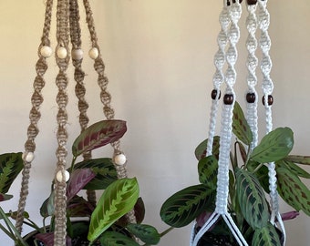 May include: Two macrame plant hangers with wooden beads. One is made of natural jute rope and the other is white cotton rope. Both are holding potted plants.