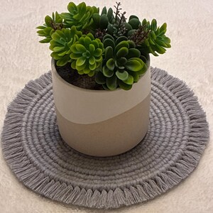 May include: A small, round, gray macrame placemat with fringe surrounds a two-tone ceramic pot. The pot holds a variety of green and brown artificial succulents. The pot is beige with a white band.