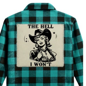 May include: A turquoise and black plaid flannel shirt with a patch featuring a cowgirl illustration and the text "THE HELL I WON'T". The patch is a light beige color with a stitched border.
