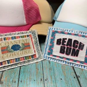 May include: A collection of trucker hats with white mesh backs and various colored fronts. Two patches are visible, one with "COASTAL COWGIRL" and a seashell design, the other with "BEACH BUM" in black letters. The hats and patches are on a weathered, turquoise wooden surface.