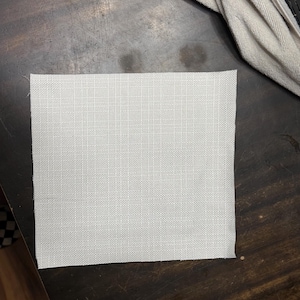 May include: A square, light gray fabric swatch with a woven texture. The fabric is placed on a dark brown wooden surface, with a beige fabric partially visible in the background.