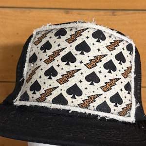 May include: Black trucker hat with a white patch featuring a pattern of black spades, orange leopard print lightning bolts, and small white stars. The hat has a black brim and a mesh back.