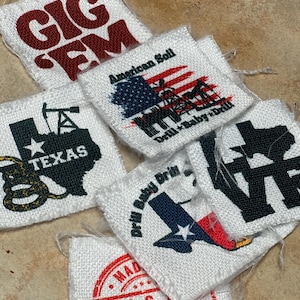 May include: A collection of white fabric patches with various designs and text. One patch reads "GIG FM" in maroon, another features the American flag with "American Soil" and "Drill + Baby + Drill". Other patches include "TEXAS" and "Drill Baby Drill".