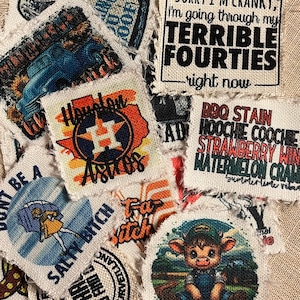 May include: A collection of fabric patches with various designs, including a cartoon cow, a baseball team logo, and a humorous saying about turning 40. The patches are made of a natural canvas material with frayed edges.