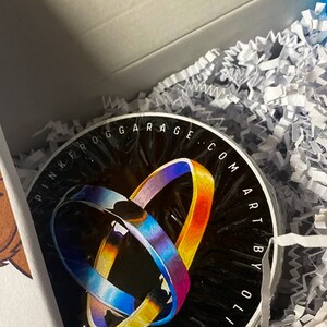 May include: A black circular sticker with the text "Pink Frog Garage: com Art by Oli" in white lettering. The sticker features a colorful abstract design of two overlapping rings.
