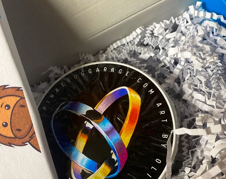 May include: A black circular sticker with the text "Pink Frog Garage: com Art by Oli" in white lettering. The sticker features a colorful abstract design of two overlapping rings.