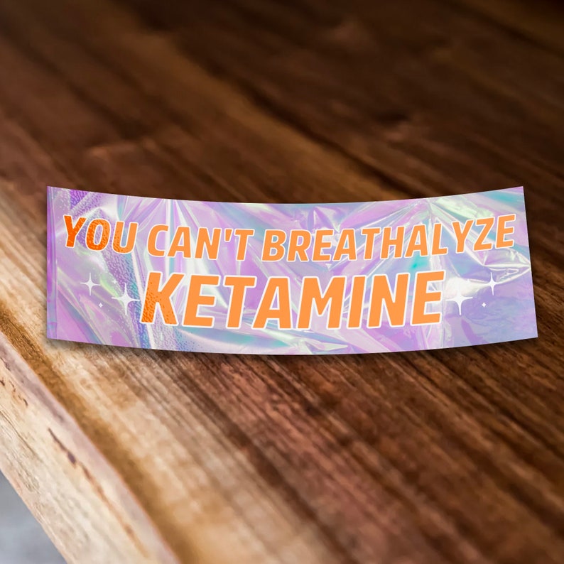 Puede incluir: Una pegatina hologr&aacute;fica con el texto "YOU CAN'T BREATHALYZE KETAMINE" en letras naranjas sobre un fondo morado con detalles de purpurina.