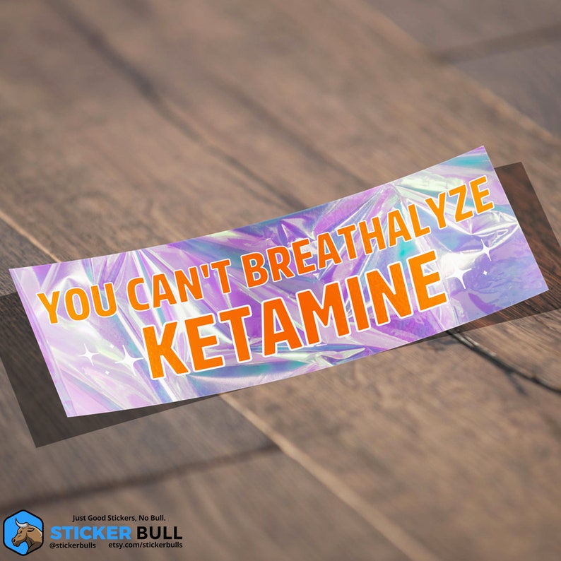 Puede incluir: Una pegatina hologr&aacute;fica con el texto "YOU CAN'T BREATHALYZE KETAMINE" en letras naranjas sobre un fondo iridiscente morado y azul.