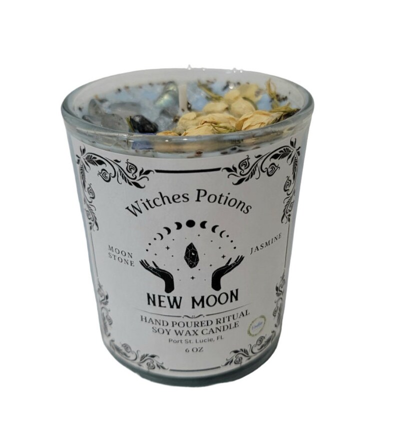 May include: A white candle jar with a black label that reads "Witches Potions New Moon Hand Poured Ritual Soy Wax Candle Port St. Lucie, FL 6 OZ". The label also features a moon and stars design. The candle jar is filled with a blue wax and contains crystals and dried flowers.