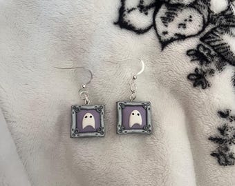 May include: A pair of handmade ghost earrings. Each earring features a white ghost design on a lavender background, framed in a gray, ornate border. The earrings have silver-colored ear wires.