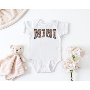 May include: White baby onesie with the word "MINI" in a leopard print design. A teddy bear and a blanket are next to the onesie. A bouquet of white flowers is also in the image.