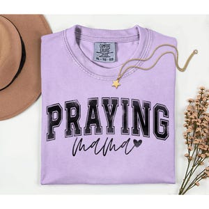 May include: A light purple t-shirt with the words "PRAYING mama" in black lettering. A gold necklace with a star pendant and a brown hat are next to the shirt. A sprig of dried flowers is on the right.
