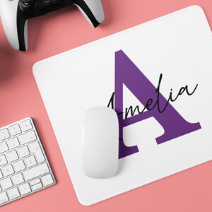 以下が含まれることがあります： 紫色の文字「A」と筆記体で「Amelia」と書かれた白いマウスパッド。白いマウスがパッドの上にあります。キーボード、ヘッドフォン、ゲームコントローラーも表示されています。