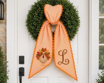 May include: A green boxwood wreath adorned with a hanging orange and white striped fabric decoration. The fabric features a scalloped orange edge, an embroidered turkey on the left tail, and a large brown embroidered letter L on the right tail.