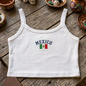 May include: White ribbed tank top with MEXICO embroidered in blue, and a small Mexican flag. The top has thin straps and a scoop neckline. The design is centered on the chest.