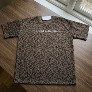 May include: A brown and black leopard print t-shirt with short sleeves and a crew neckline. The text "i need a die coke." is printed in white lowercase letters across the center of the chest. A small white rectangular tag is attached at the neck.