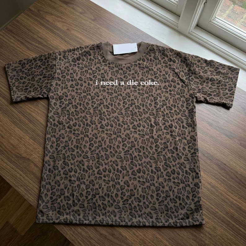 May include: A brown and black leopard print t-shirt with short sleeves and a crew neckline. The text "i need a die coke." is printed in white lowercase letters across the center of the chest. A small white rectangular tag is attached at the neck.